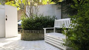 A white bench in a garden area with a paved walkway, surrounded by greenery and a white wall.