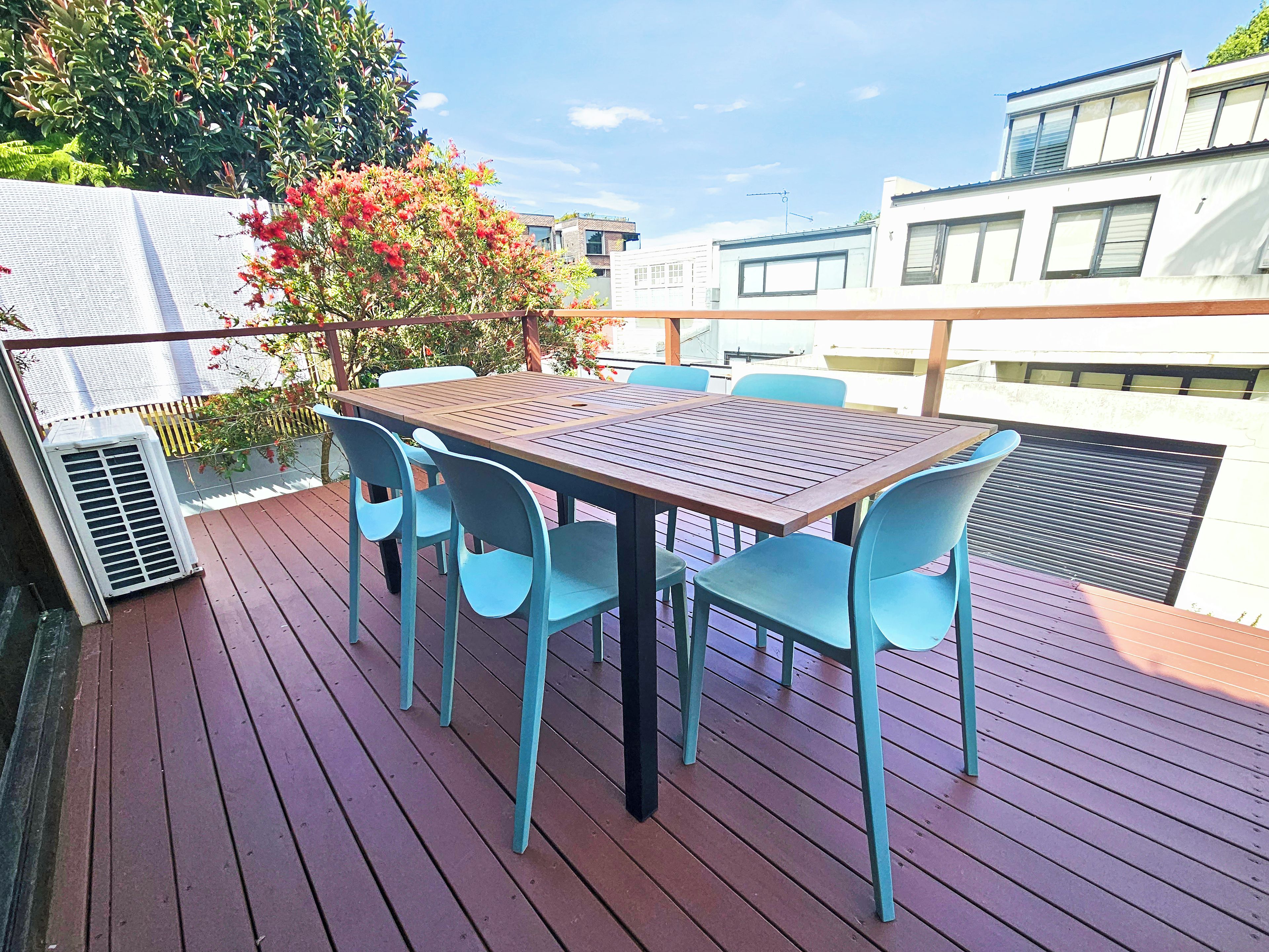 Une terrasse extérieure avec une table en bois et des chaises bleues, entourée de verdure et de bâtiments.