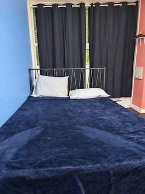A bed with a dark blue bedspread and white pillows, a metal headboard, and a window with black curtains.