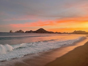 Una spiaggia con onde, un tramonto e formazioni rocciose in lontananza.