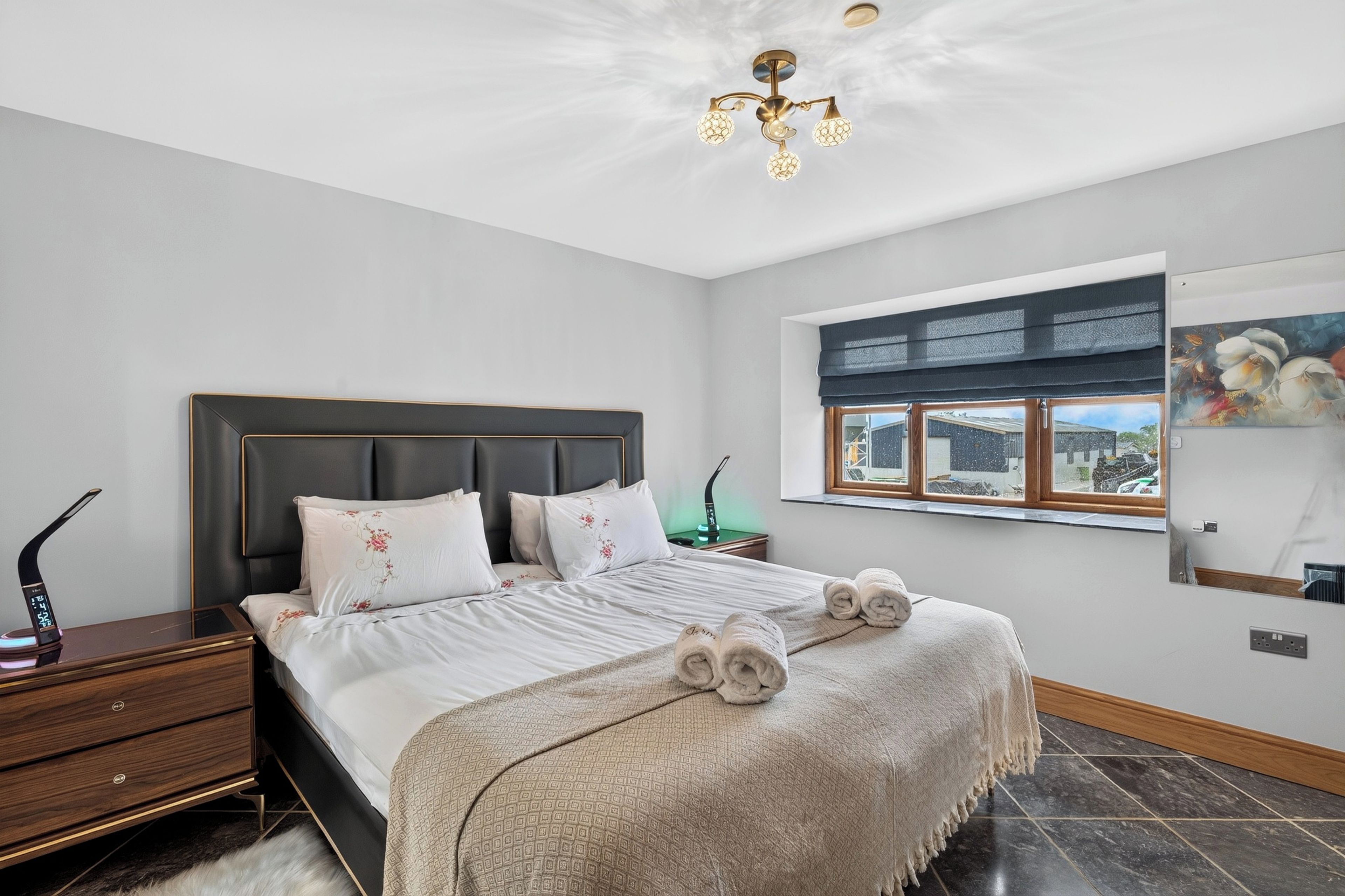 A bedroom with a bed, a nightstand, a lamp, a window with a view, and a painting on the wall.