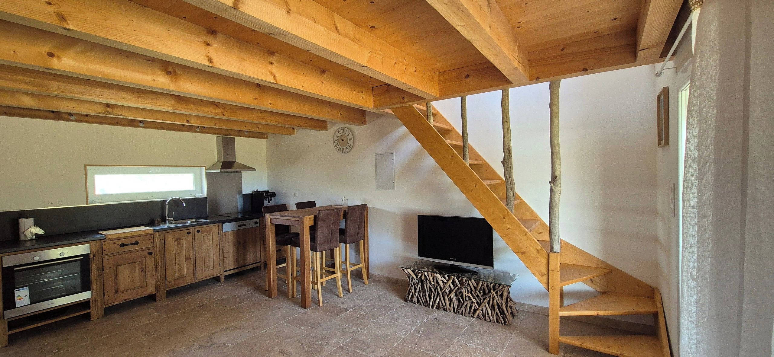 Una cocina rústica con vigas de madera, un comedor con sillas, una escalera y un televisor.