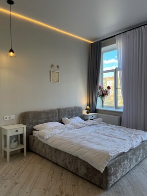 Ein Schlafzimmer mit einem Bett, Nachttischen, einem Fenster mit Vorhängen, einer Wandleuchte und einem gerahmten Bild.