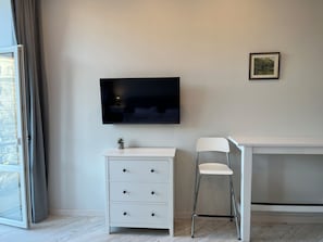 A room with a flat-screen TV mounted on the wall, a white desk with a chair, a small plant on the desk, and a window with curtains. - Beautiful apt. on Svobody avenue 7 Lviv (L'viv)