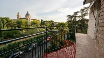 A balcony with a metal railing overlooking a street with parked cars and a building with a dome in the background.