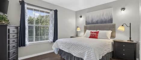 A bedroom with a bed, a nightstand, a dresser, a window with curtains, and a painting on the wall.