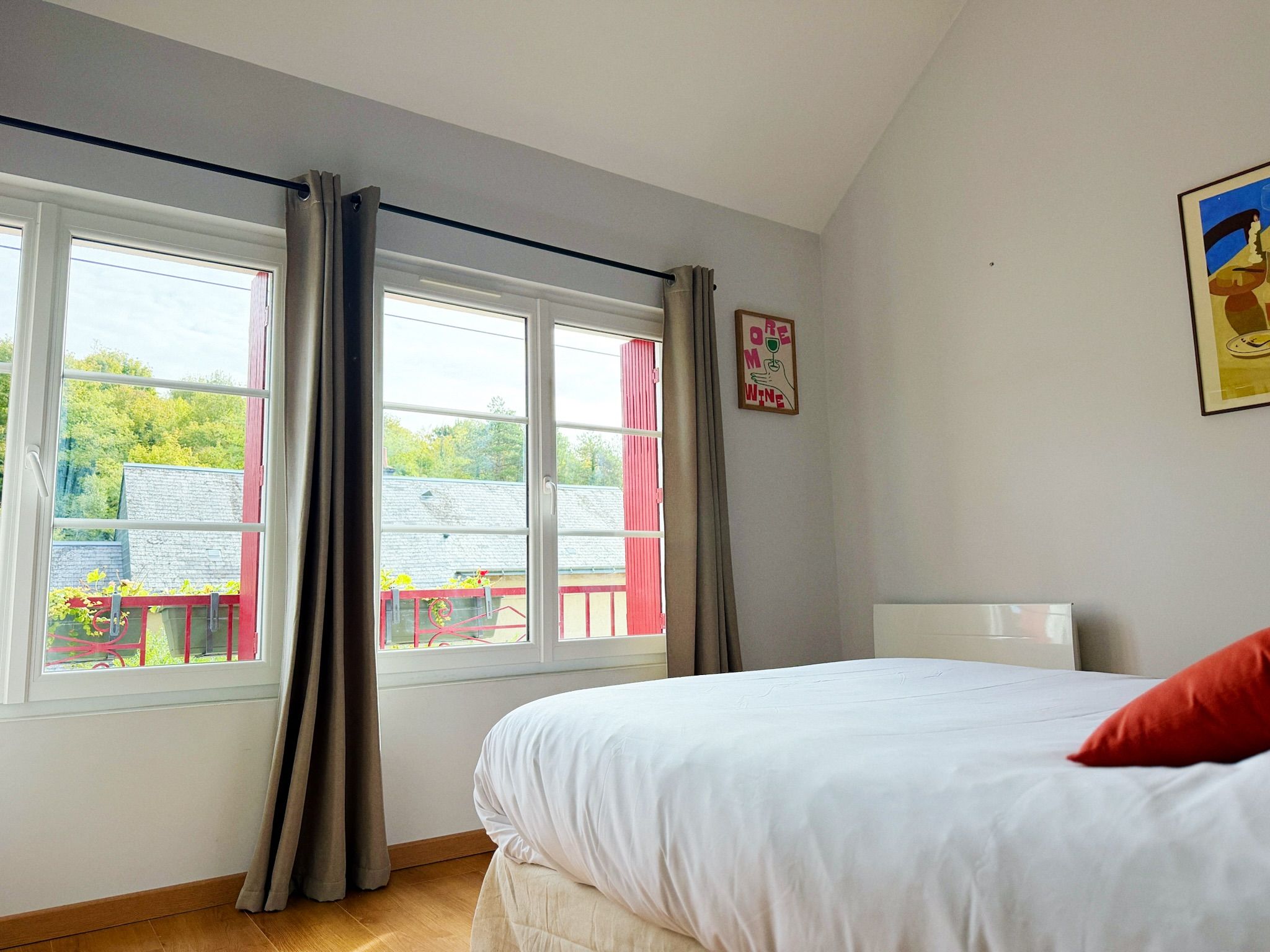Une chambre à coucher avec un lit, deux fenêtres avec des rideaux, une vue sur un bâtiment rouge à l’extérieur, et un tableau encadré au mur.