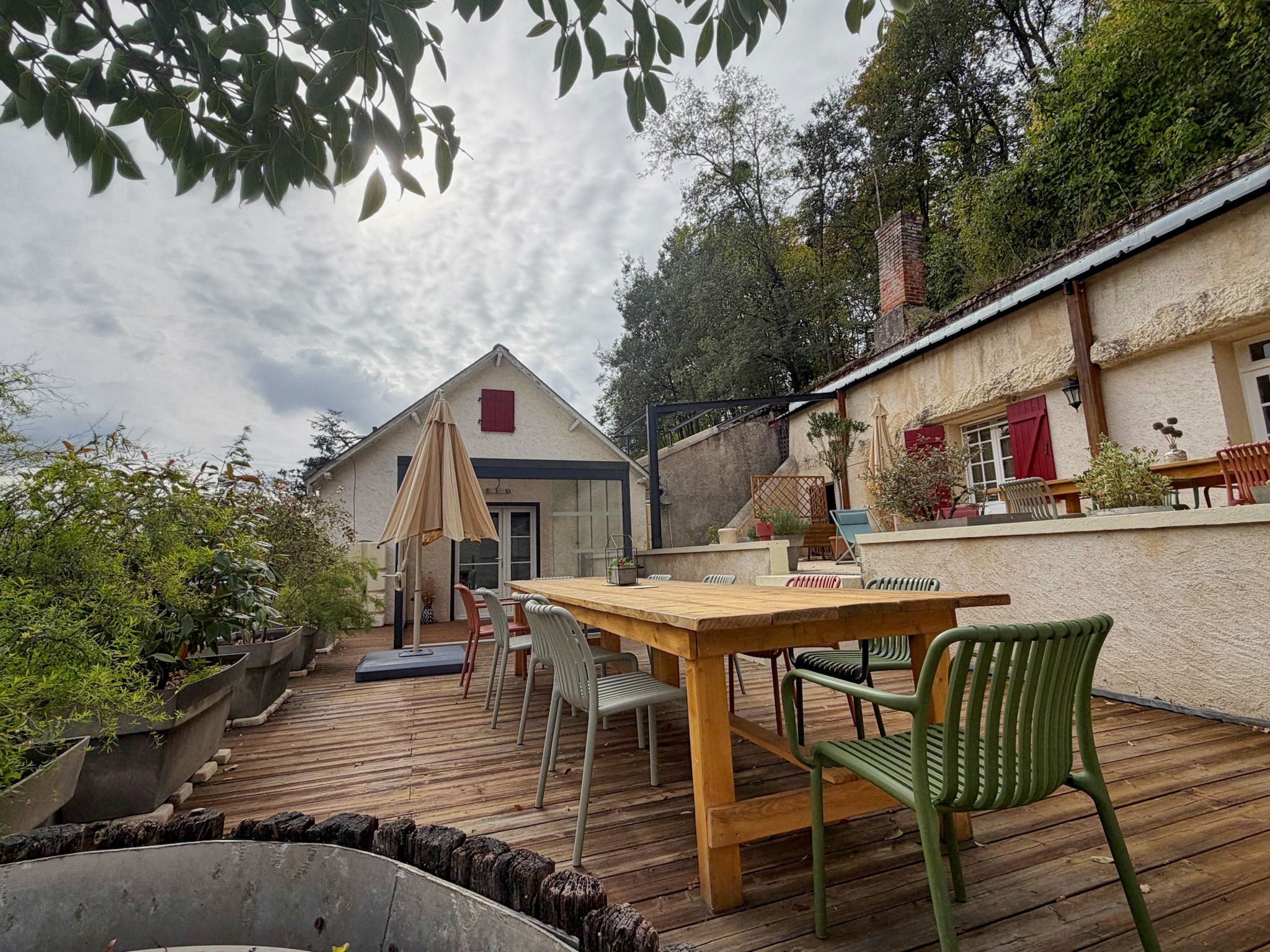 Une terrasse extérieure avec une table et des chaises en bois, entourée de plantes en pot et d’un mur de briques.