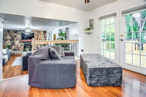 A living room with a stone fireplace, a grey sofa, and a large window with a view of the outdoors. - Hot Tub Haven with Game Room Home with In-Law Suite (Rochester)