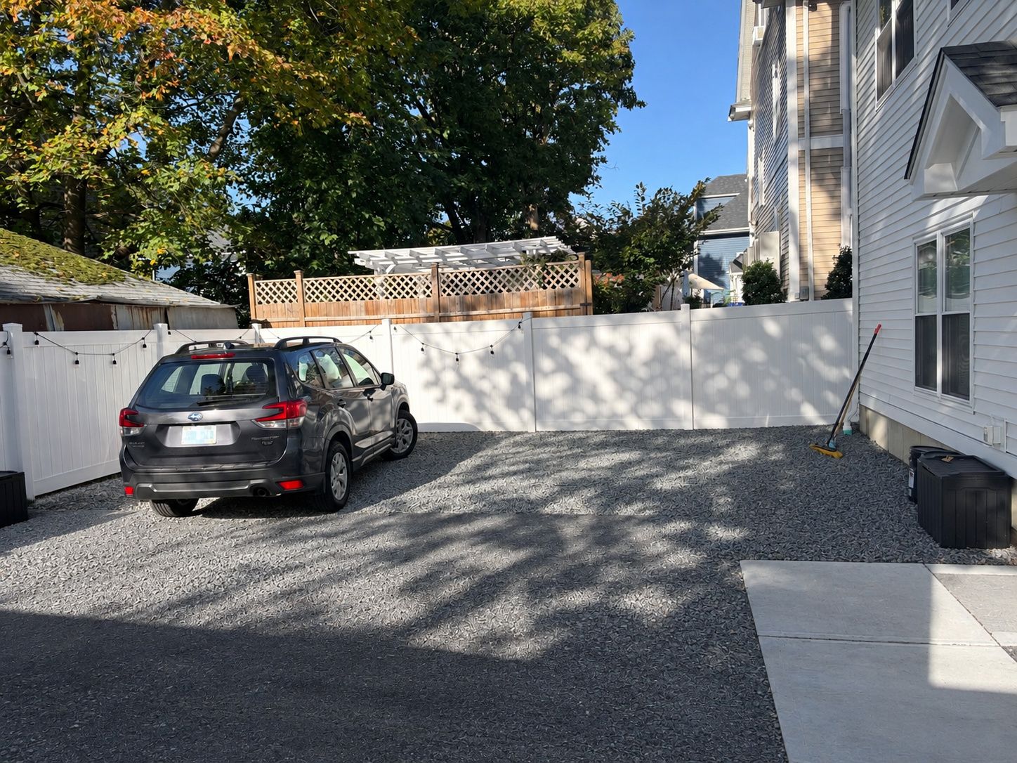 A residential driveway with a car parked, a white fence, and a building in the background.
