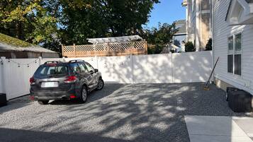 A residential driveway with a car parked, a white fence, and a building in the background.