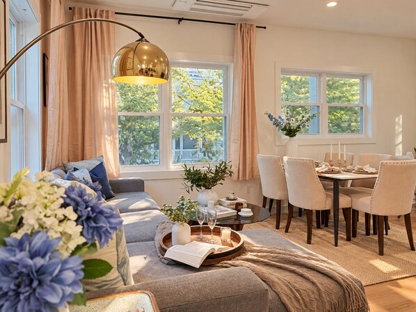 A well-lit living room with a sofa, dining table, and chairs. There are windows with curtains, a floor lamp, and a tray with a book and glasses.