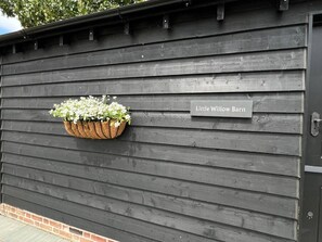 Una pared de madera negra con una maceta colgante de flores y una placa que dice 'Little Willow Barn'.