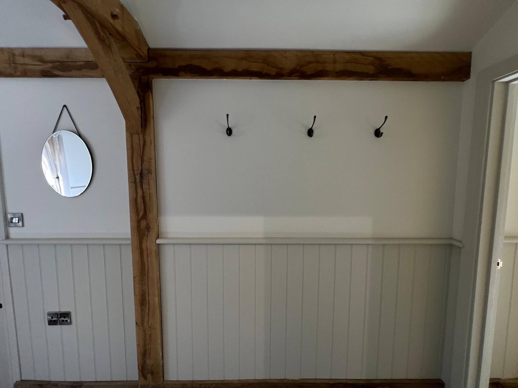 A room with a white paneled wall, a wooden ceiling beam, and three wall-mounted hooks.