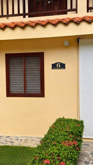 A building with a number sign indicating its unit, a window with shutters, a small garden with green shrubs and pink flowers, and a tiled roof. - Tremendous 3-bedroom plus one bed loft condo with AC in Las Terrenas (Las Terrenas)