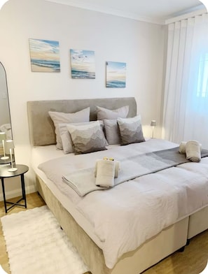 A neatly made bed with pillows and towels, a bedside table with a lamp, and a view of the outdoors through a window. - Enjoyable 1-bedroom apartment in vibrant Bloubergstrand (Cape Town)