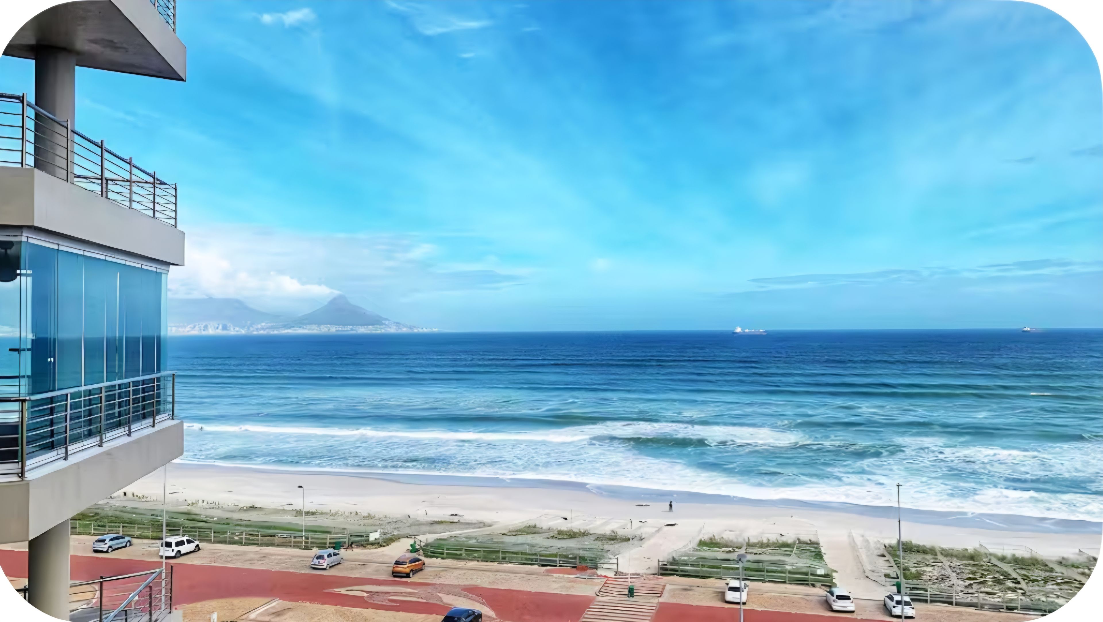 A coastal view with a building overlooking the beach and ocean, a distant mountain, and a ship in the distance.