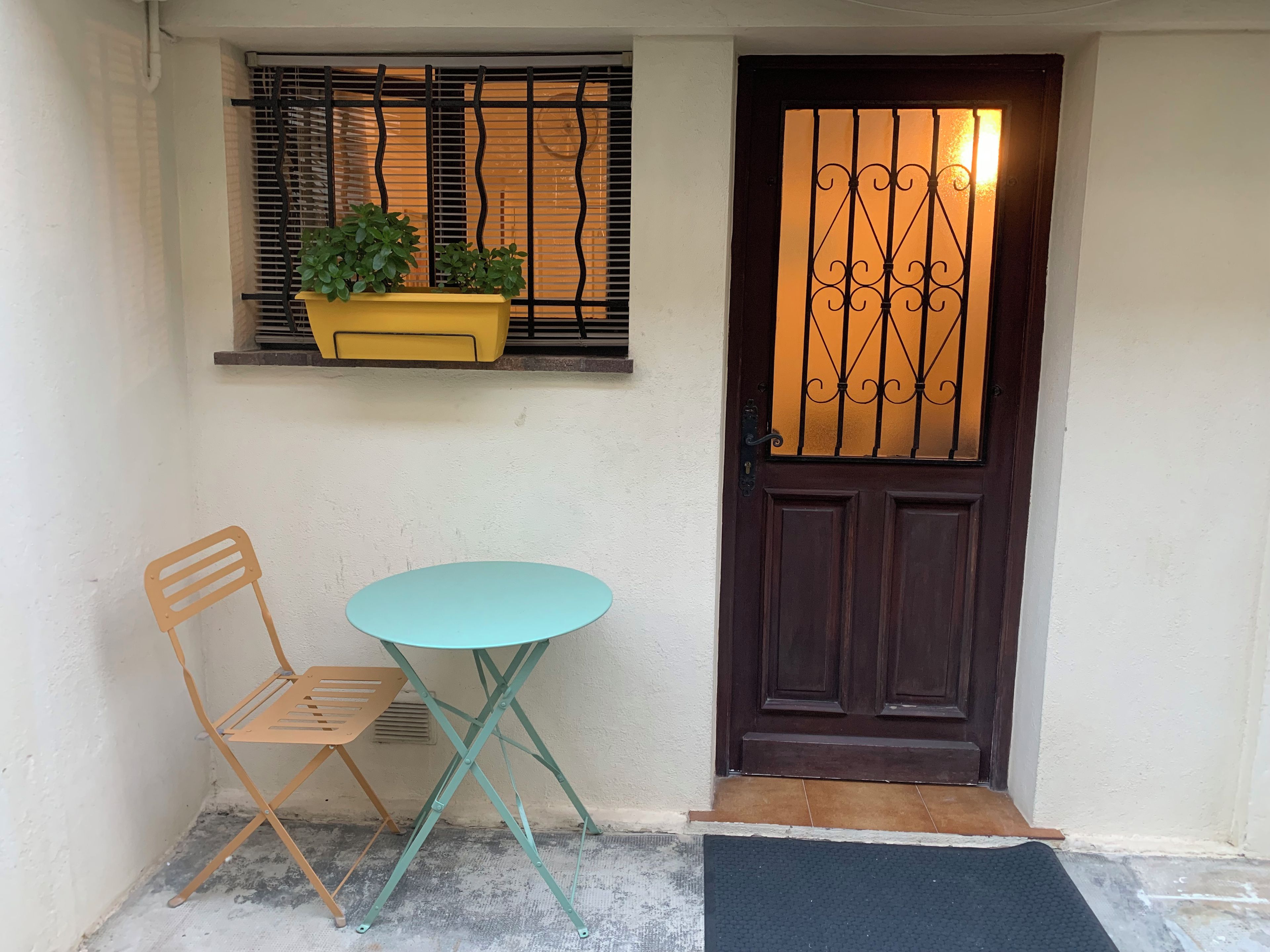 A small outdoor seating area with a table and a chair, a door with a decorative metal grille, and a window with a plant.