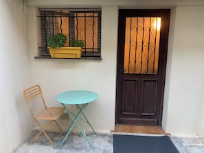 Ein kleiner Sitzbereich im Freien mit Tisch und Stuhl, eine Tür mit dekorativem Metallgitter und ein Fenster mit Pflanze.