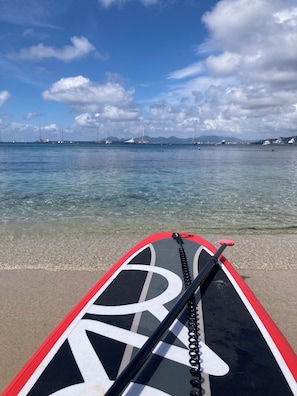 En SUP-bräda (ståbräda) med ett svartvitt mönster, ett rep och en paddel, placerad på en sandstrand med klart vatten och båtar i bakgrunden.