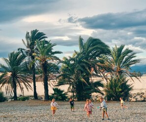 Eine Gruppe Kinder spielt an einem Kiesstrand mit Palmen und einem bewölkten Himmel.