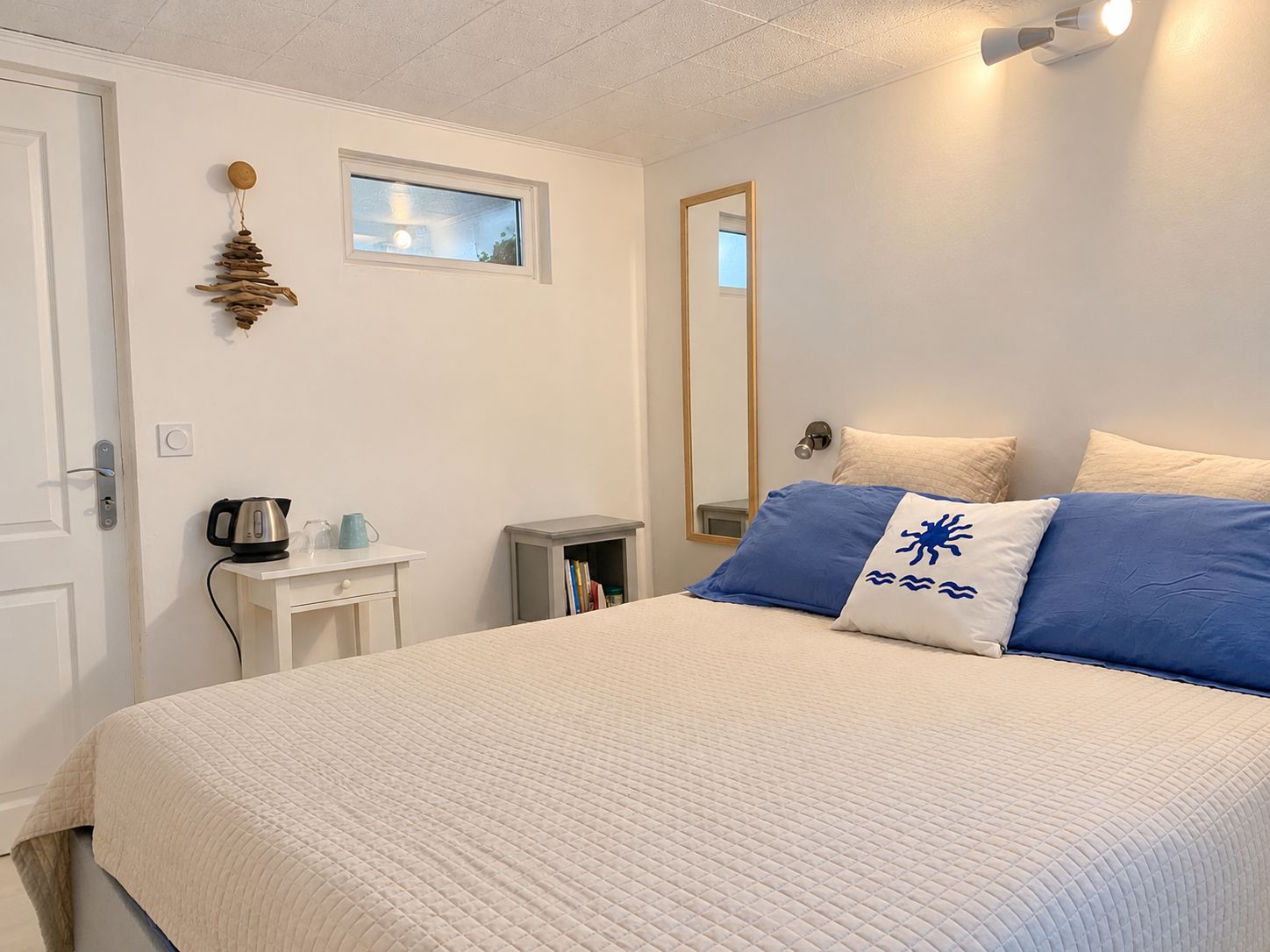 A bedroom with a bed, a bedside table with a kettle, a small shelf, a window, and a mirror on the wall.