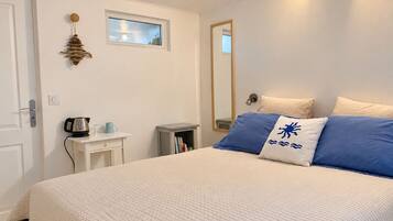 A bedroom with a bed, a bedside table with a kettle, a small shelf, a window, and a mirror on the wall.