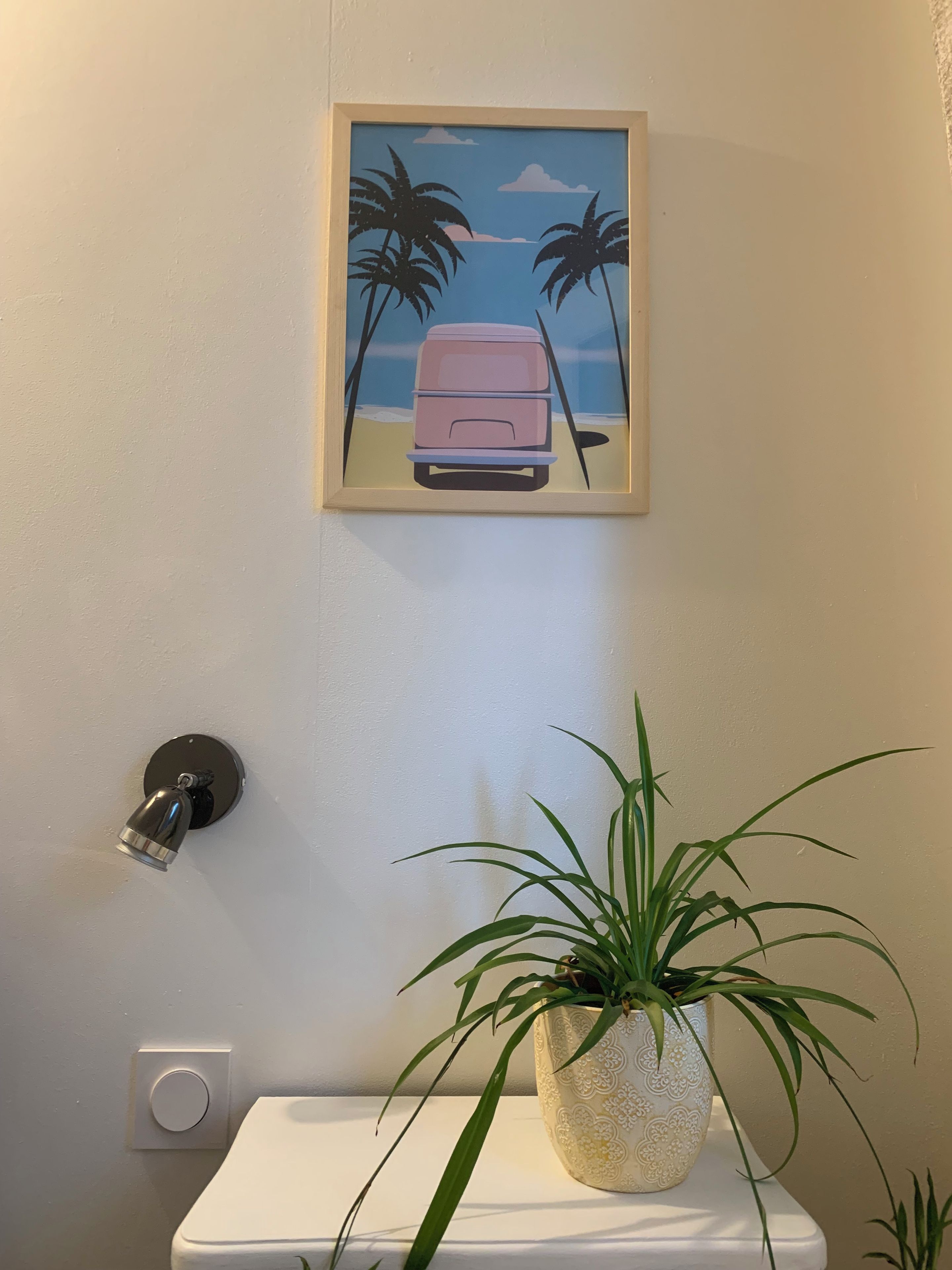 A framed artwork of a pink van on a wall, a wall-mounted light fixture, and a potted plant on a white surface.