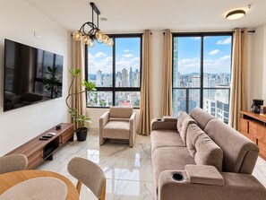 A modern living room with a flat-screen TV, a sofa, a chair, a coffee machine, and a view of the cityscape through large windows. - Cozy 1-bedroom apartment in perfect Panamá with AC and gym access (Panamá)