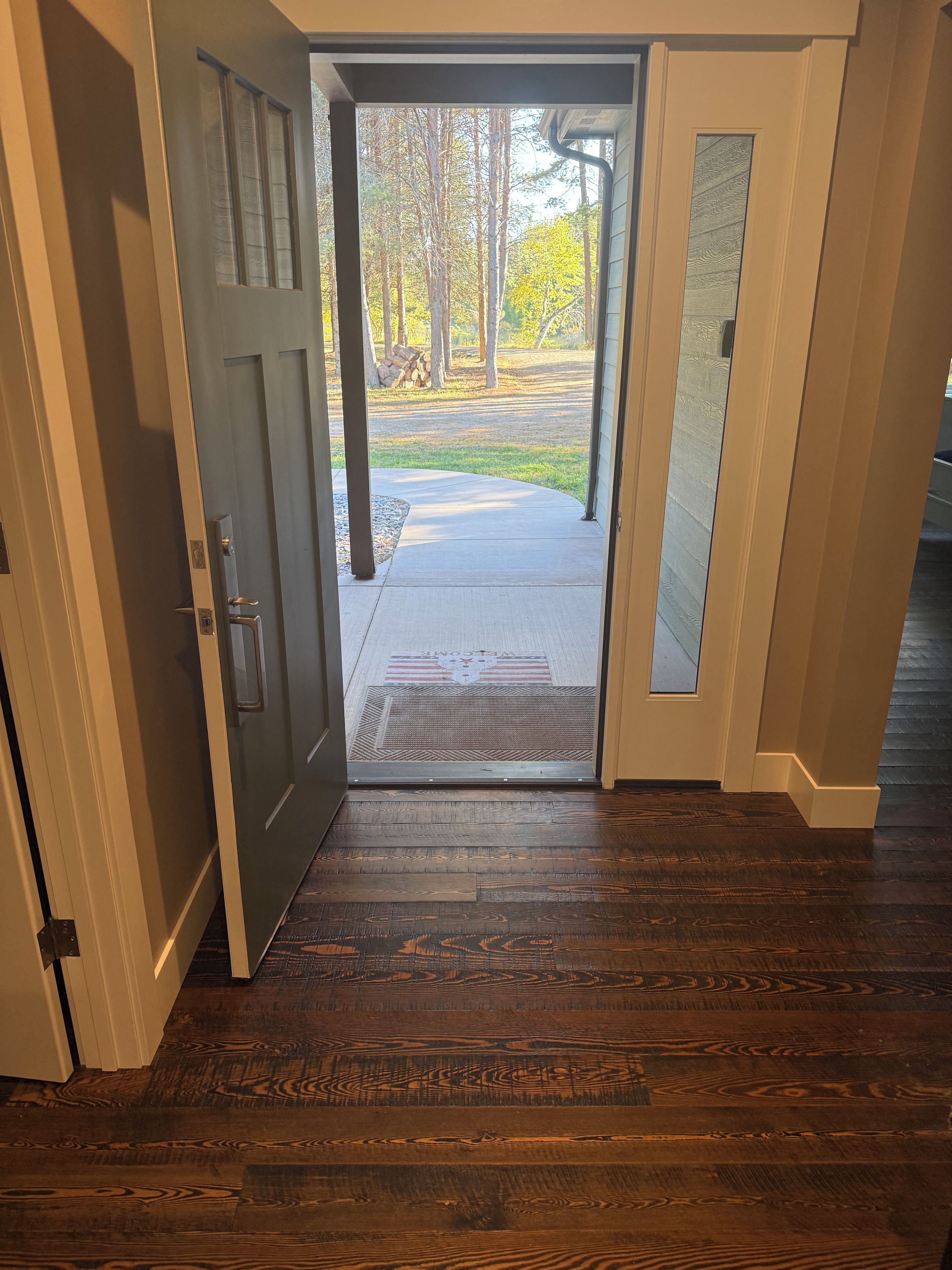 A wooden floor leading to a door with a glass panel, a welcome mat, and a view of a yard with trees and a fire pit.