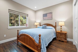 A bedroom with a wooden bed, two bedside tables with lamps, a window with a view of trees, and an American flag on the wall. - Freedom House on Swan River - 3BR/2BA (Bigfork)