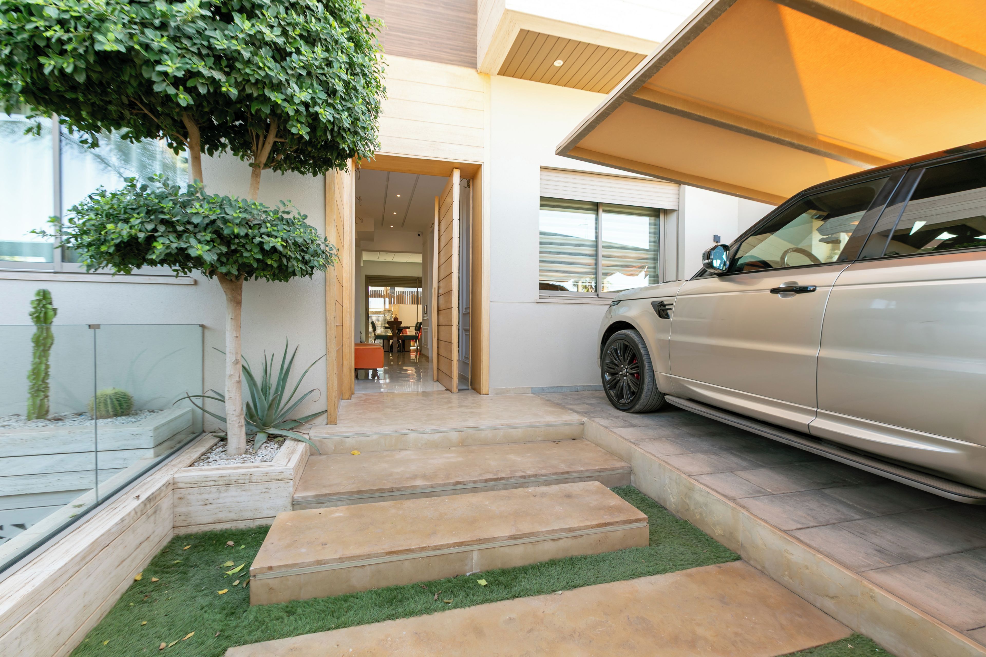 A modern house with a car parked in the driveway, a small garden with a tree and a cactus, and a clear view into the interior.