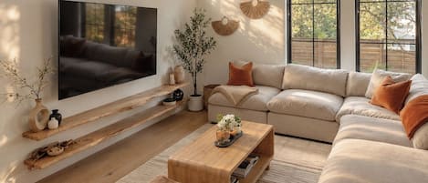 A modern living room with a large flat-screen TV, a beige sectional sofa, a wooden coffee table, and a ceiling fan.