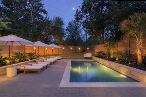 Un área de piscina con sillones de descanso y sombrillas, rodeada por una valla de madera y árboles.