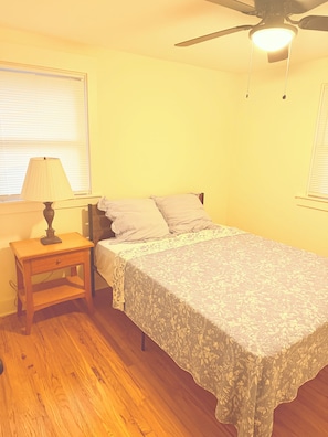 Un dormitorio con cama, mesita de noche, lámpara, ventilador de techo y ventana con persianas.