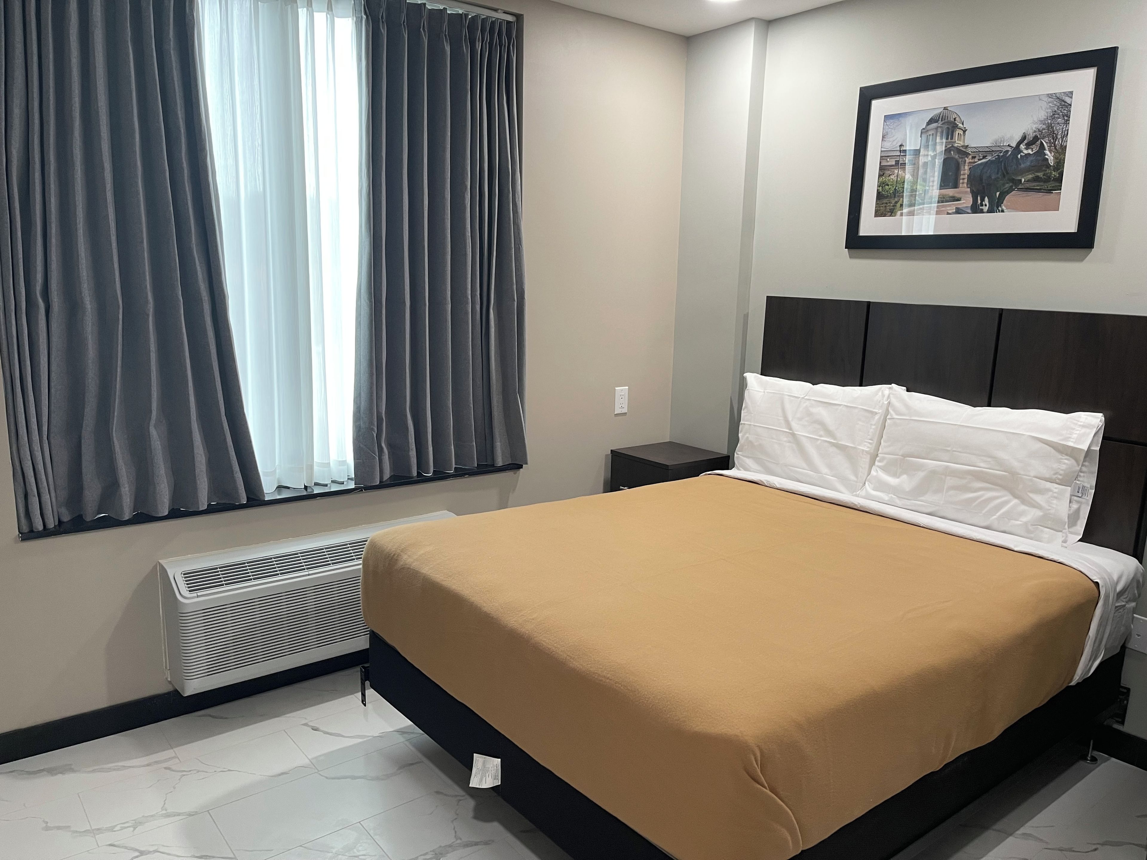 A hotel room with a bed, a nightstand, a framed picture on the wall, and a window with curtains.