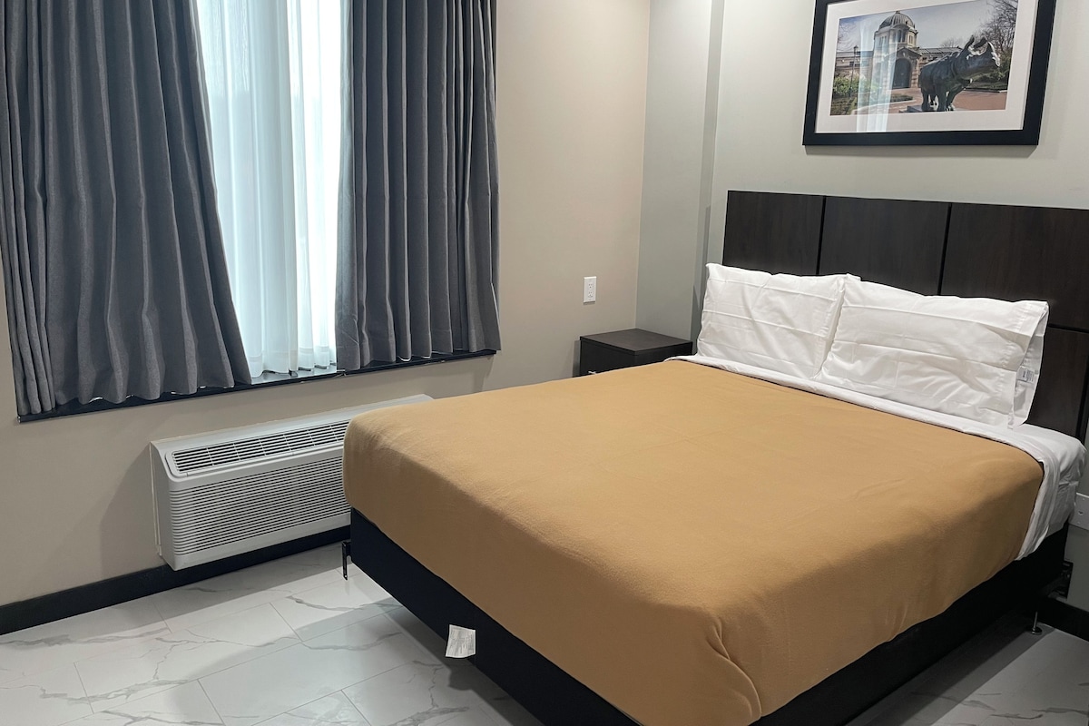 A hotel room with a bed, a nightstand, a framed picture on the wall, and a window with curtains.