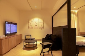 A modern hotel room with a flat-screen TV on a wooden cabinet, a round table with a chair, a sofa, and a bed with a headboard. - Chalker Residences (Aso)