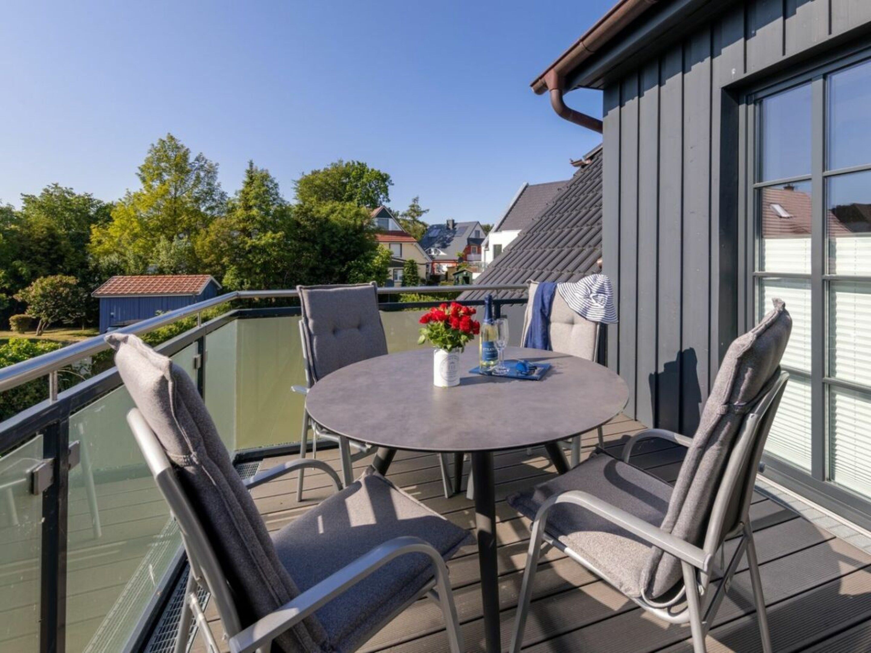 A round table with four chairs on an outdoor balcony with a glass railing, overlooking a residential area.