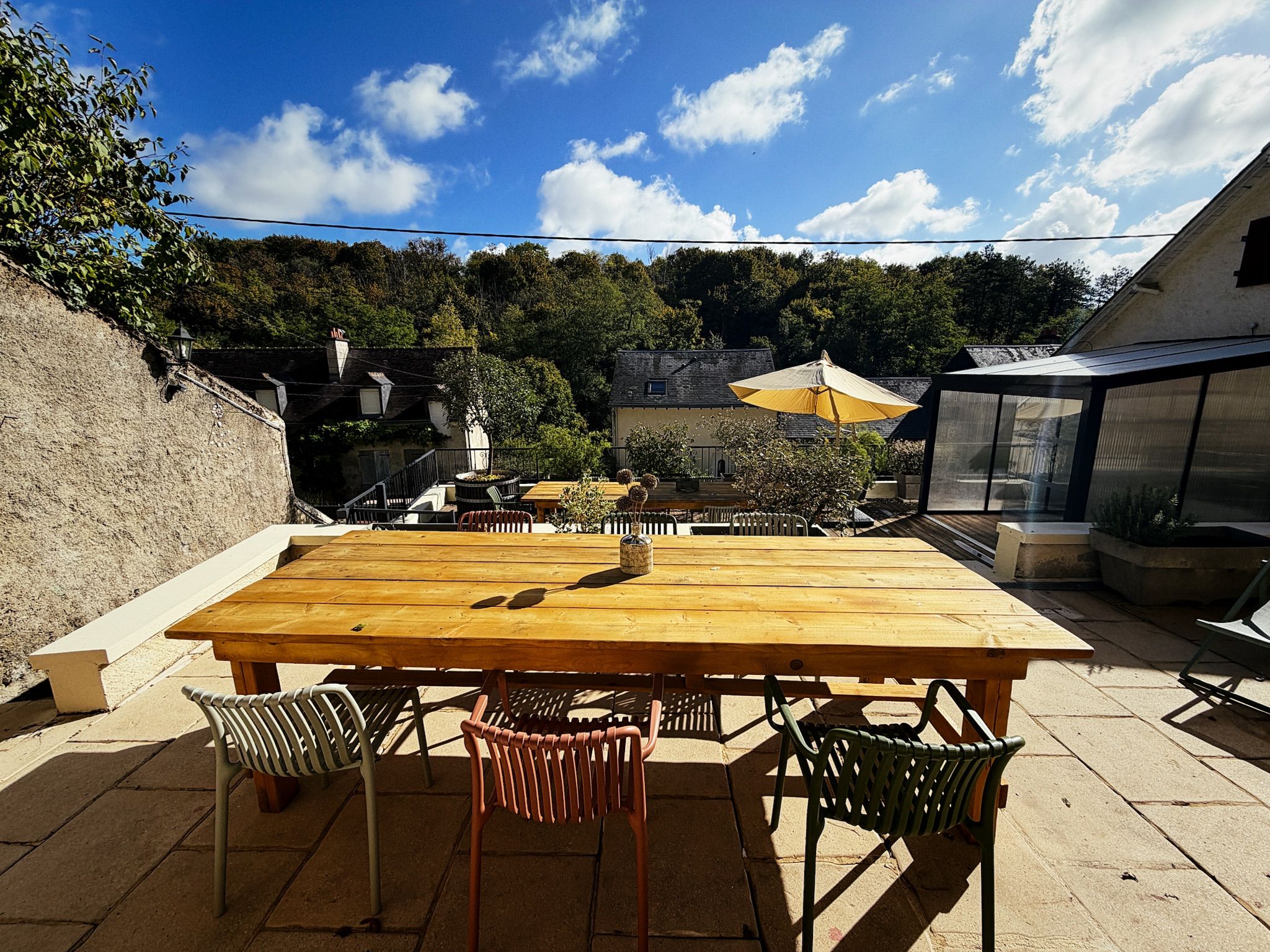 Une terrasse extérieure avec une table et des chaises en bois, entourée de verdure et de bâtiments.