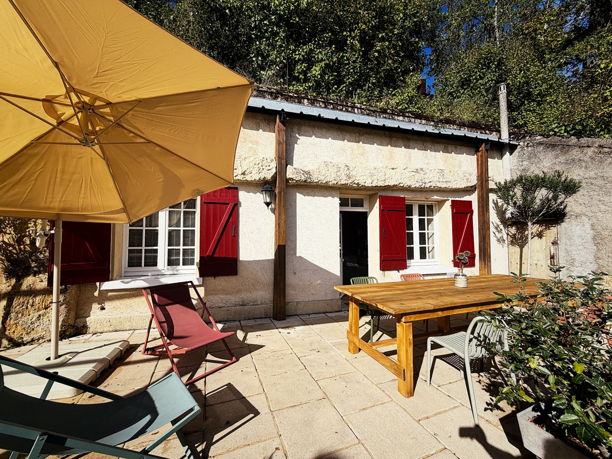 A rustic outdoor patio with a wooden table and chairs, a yellow umbrella, and a stone wall.