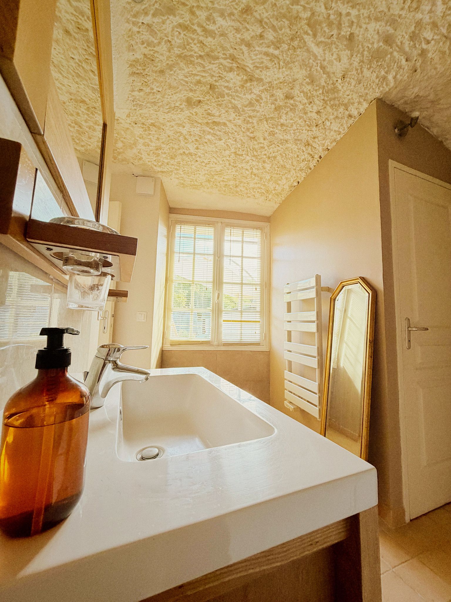 Une salle de bain avec un lavabo blanc, un miroir et une fenêtre avec des stores.