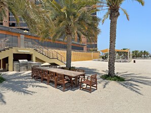A beachside outdoor seating area with a grill, wooden tables, and chairs, surrounded by palm trees and a building with a staircase. - Luxury 3BD Apartment in Palm Jumeirah | Marina Views & Stunning Sunsets (Dubai)