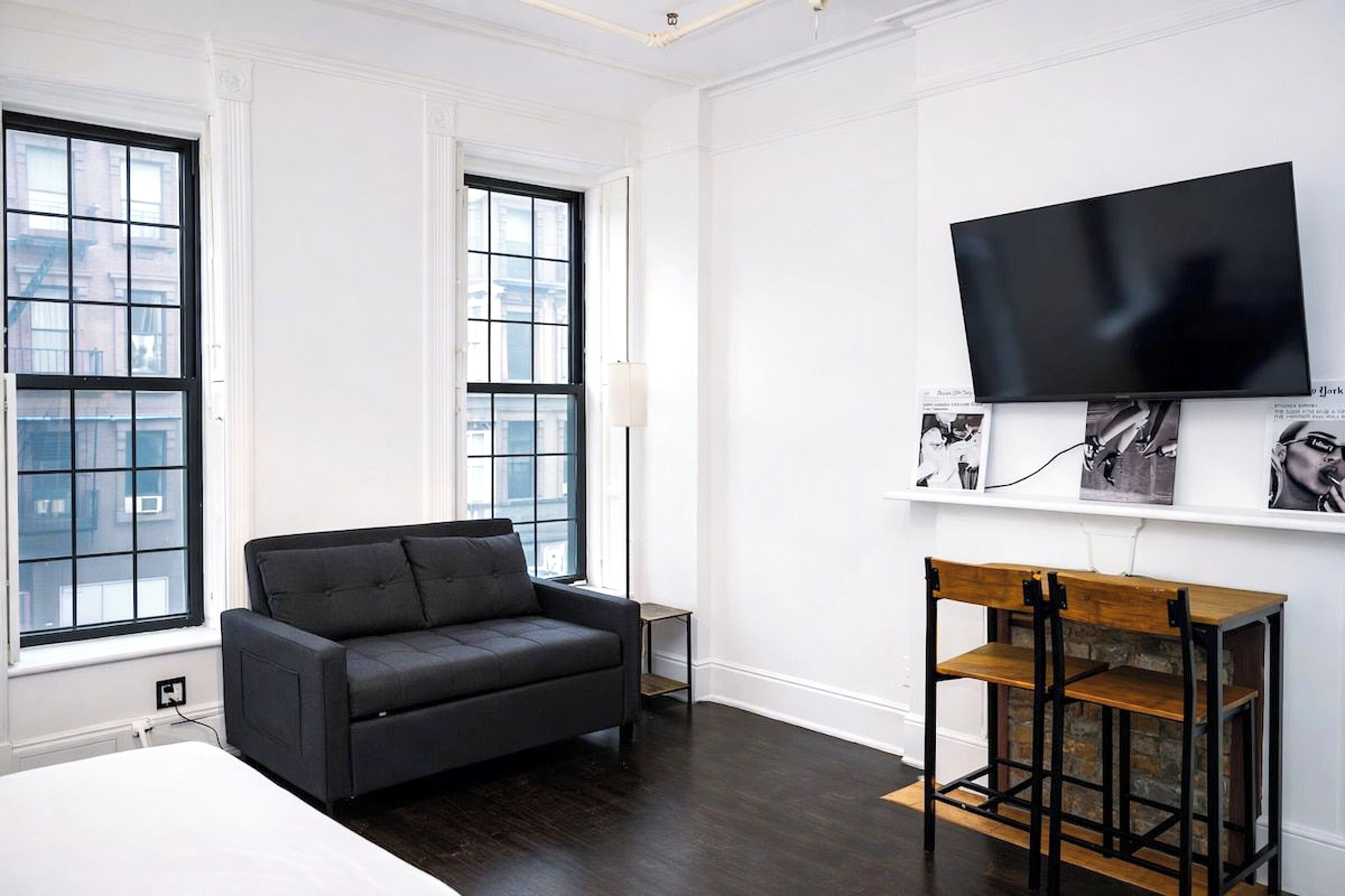 A modern hotel room with a large window, a black sofa, a wooden side table, and a mounted flat-screen TV.