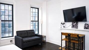 A modern hotel room with a large window, a black sofa, a wooden side table, and a mounted flat-screen TV.