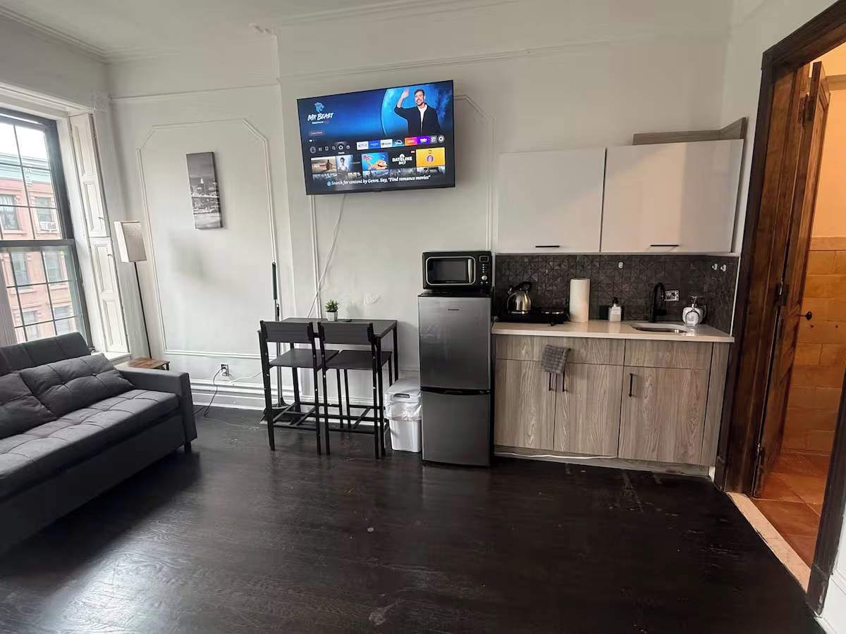 A modern living room with a sofa, a dining table with chairs, a kitchen area with a microwave, refrigerator, and cabinets, and a flat-screen TV mounted on the wall.