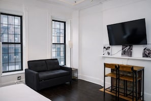 A modern hotel room with a dark wood floor, a black sofa, a wooden side table, a flat-screen TV mounted on the wall, and large windows with a city view. - Lexington studio (New York)