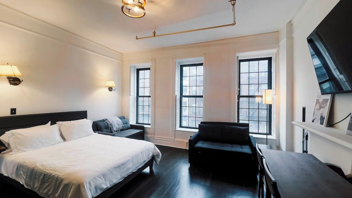 A hotel room with a large bed, a sofa, a desk, and a TV. There are windows and a ceiling light.