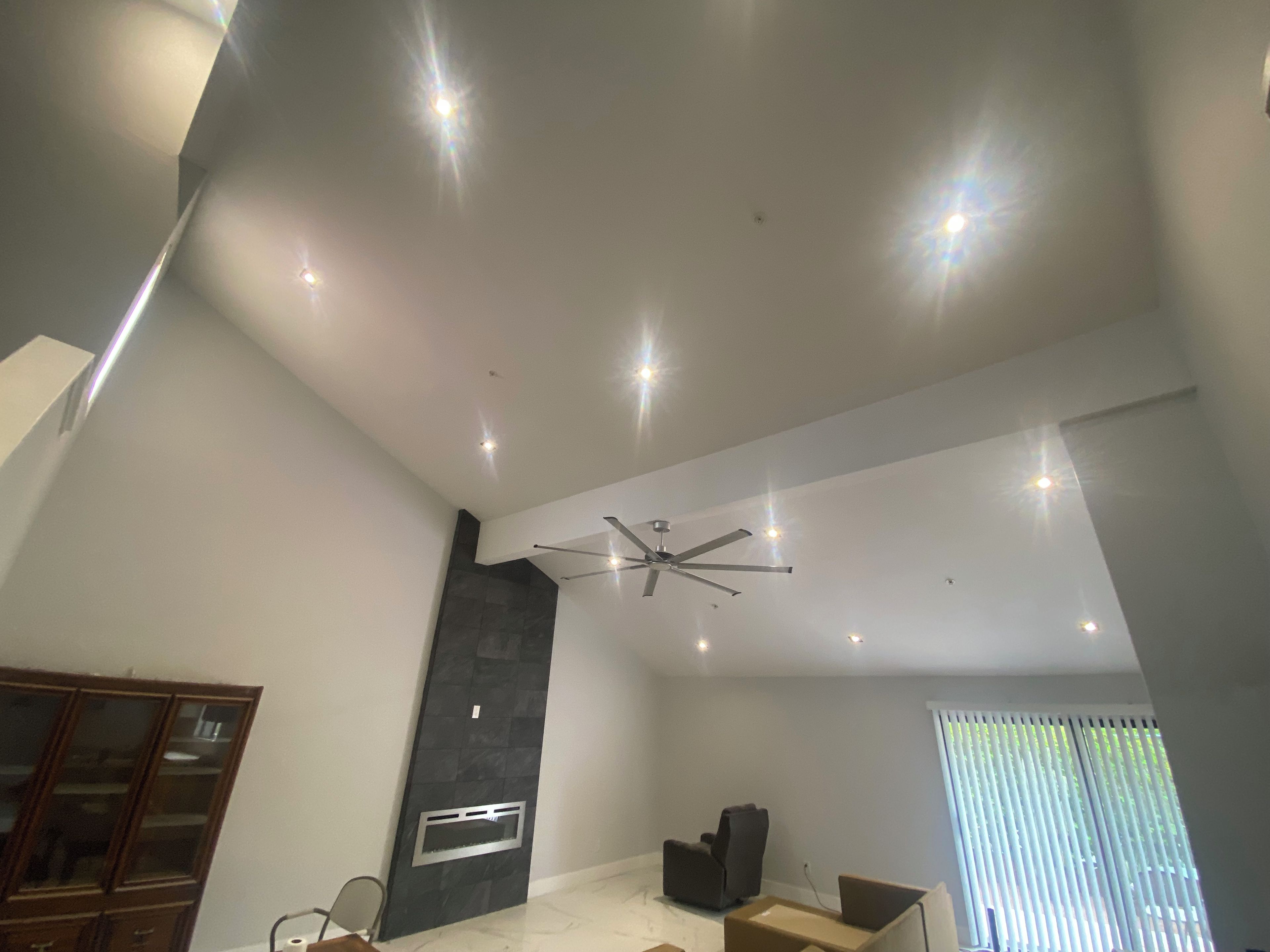 A modern living room with a high ceiling, recessed lighting, a ceiling fan, and a large window with blinds.