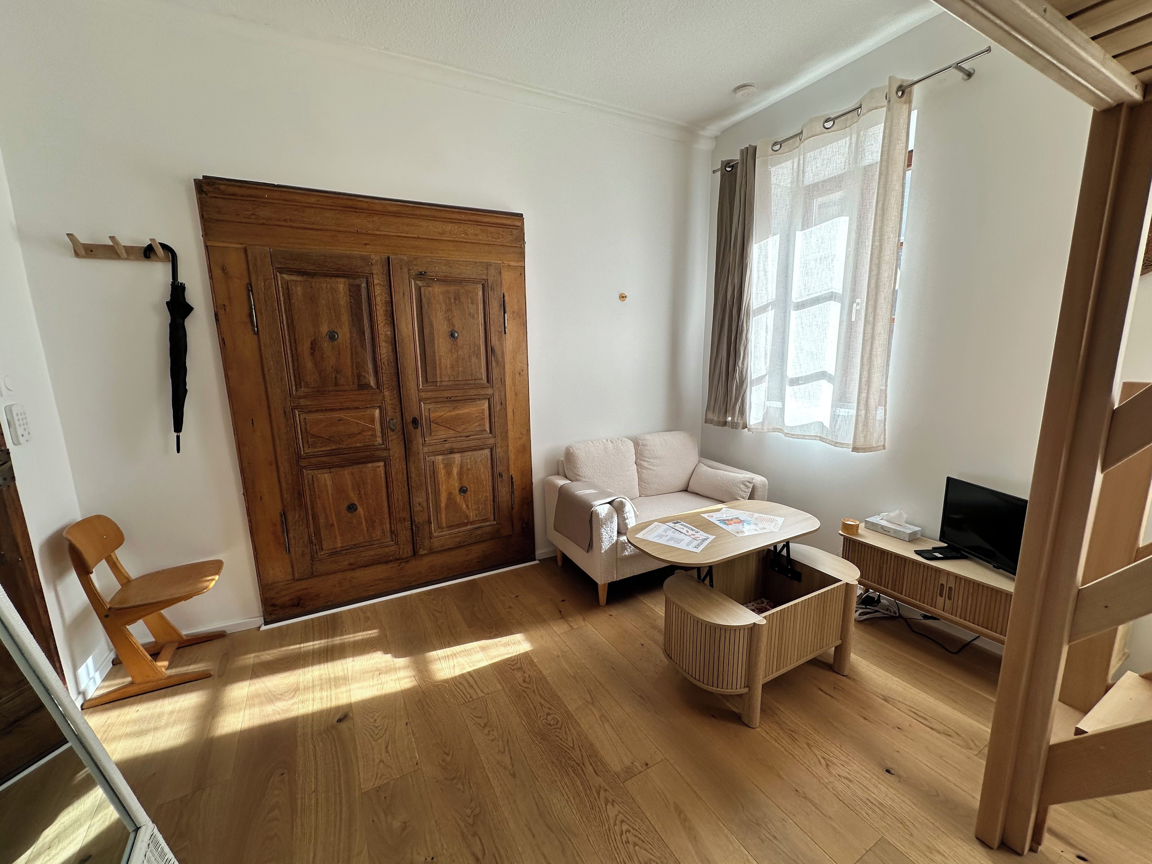 Una habitación con un armario de madera, un sofá, una mesita con papeles, un televisor y una ventana con cortinas.